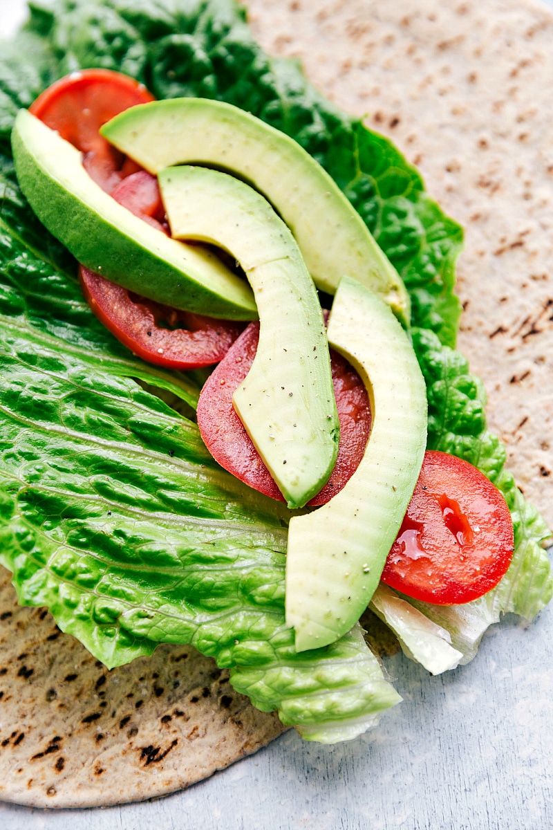 Image of the tortilla with lettuce, tomatoes, and avocado on it for Chicken Salad Wraps.