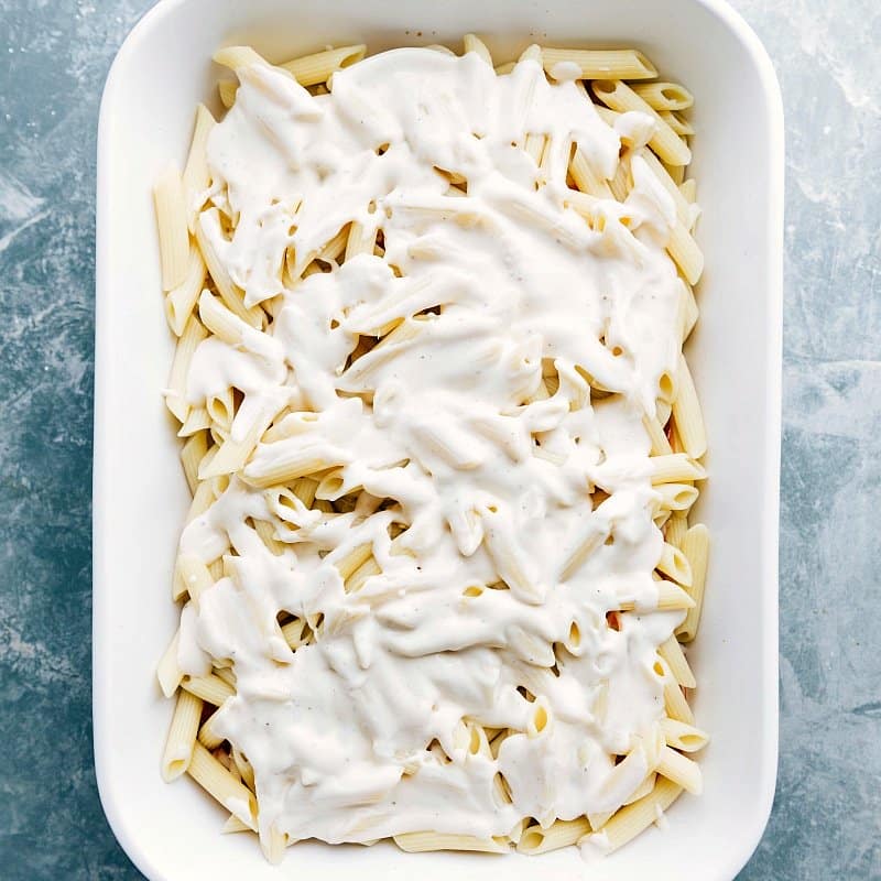 White sauce being poured over a casserole full of noodles, ready for baking.