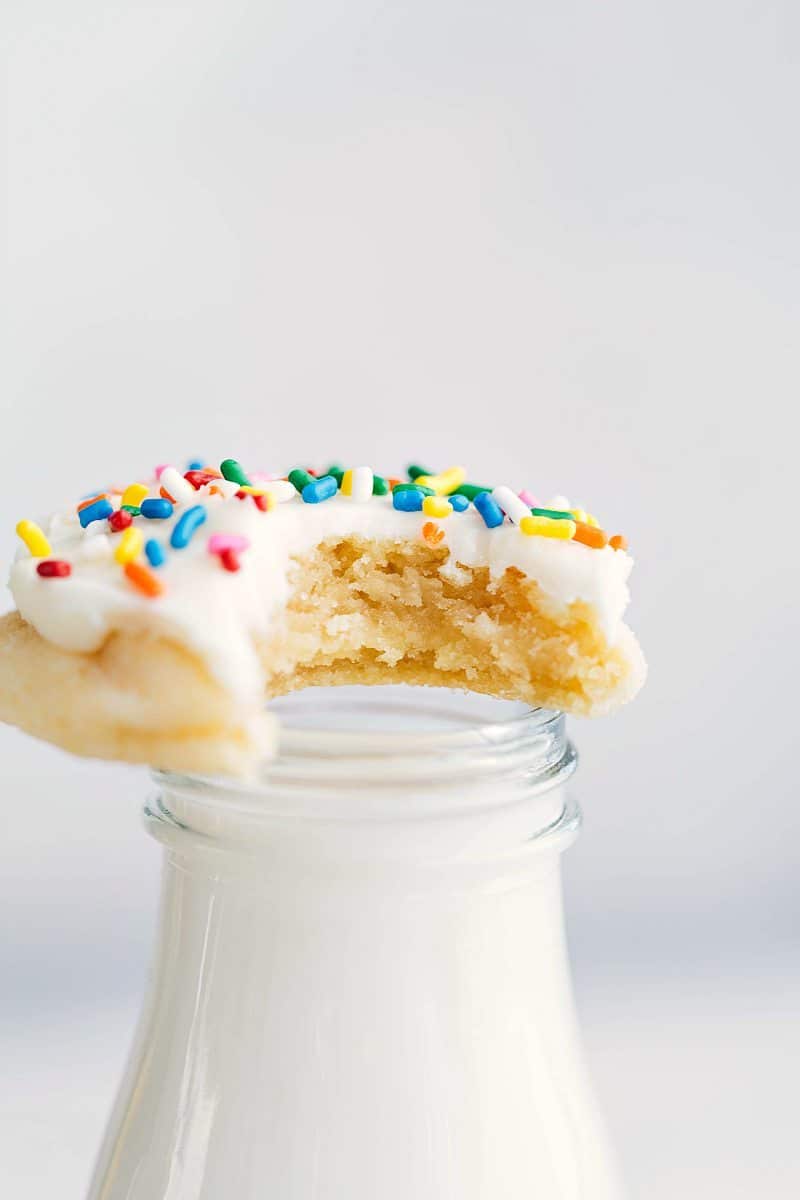 A large bite taken from a delicious soft sugar cookie topped with colorful sprinkles, resting on a container of milk.