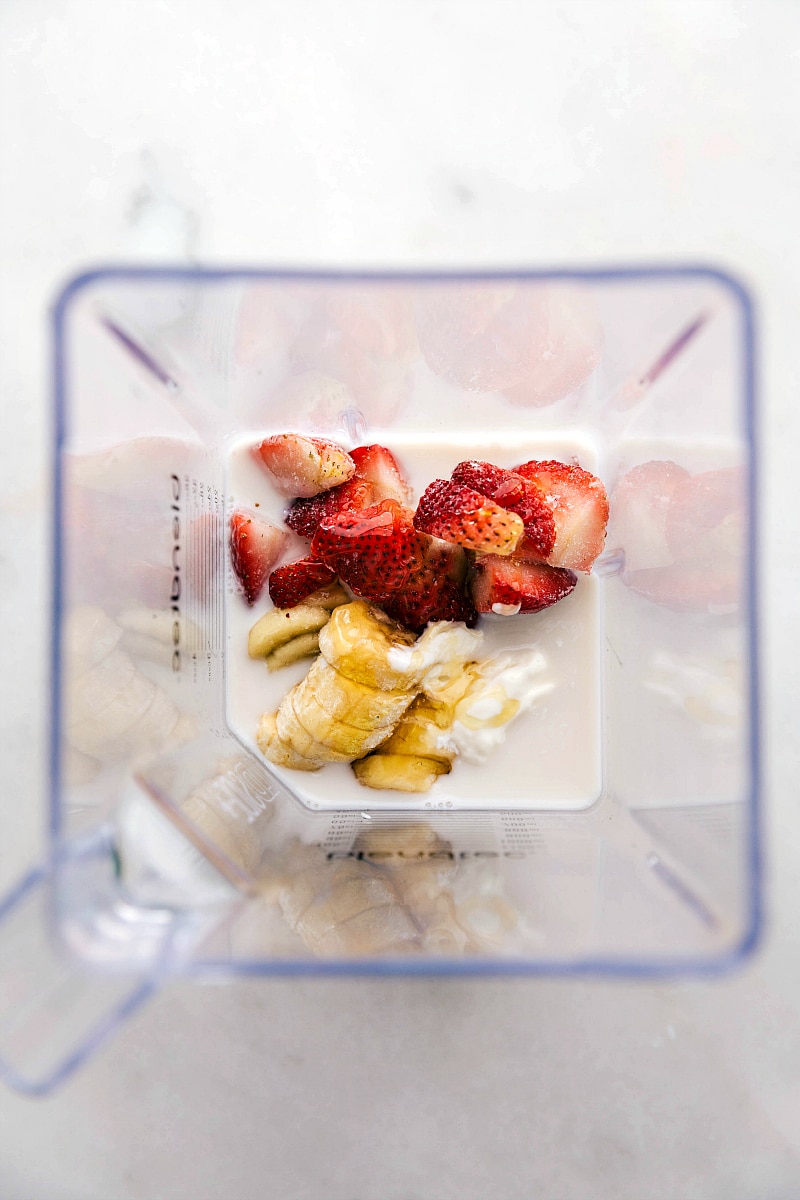 Overhead process shot of the ingredients with almond milk, Greek yogurt, bananas, honey, and strawberries.
