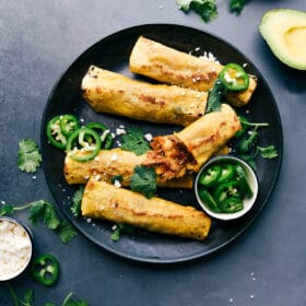 Finished crunchy pork taquitos on a plate, accompanied by sliced jalapeños, cilantro, and avocado on the side.