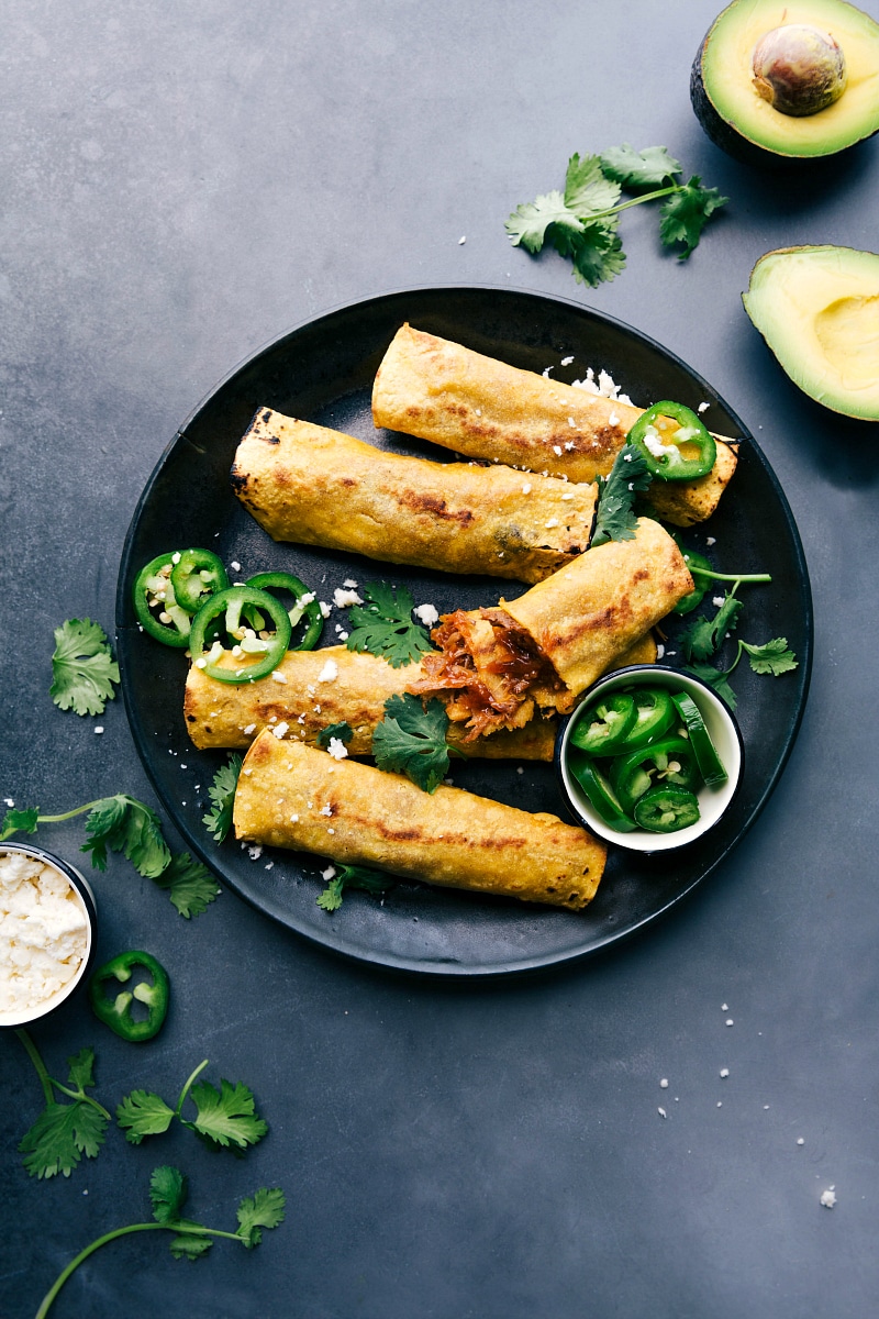 Finished crunchy pork taquitos on a plate, accompanied by sliced jalapeños, cilantro, and avocado on the side.