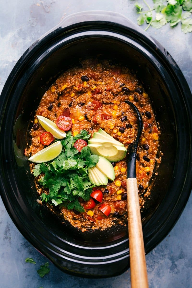 Crockpot quinoa enchilada freshly made, served with a spoon and garnished with cilantro, tomatoes, avocado slices, and lime wedges.