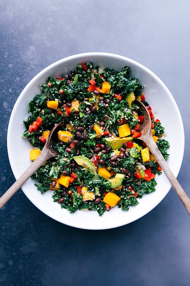 Serving bowl filled with colorful couscous kale salad, accompanied by two wooden serving spoons.