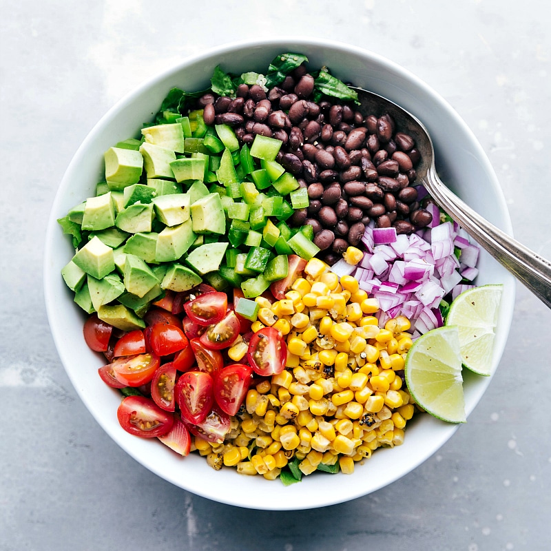 The remaining ingredients, including black beans, corn, chopped onions, tomatoes, and more, being added to the bowl of quinoa and lettuce.