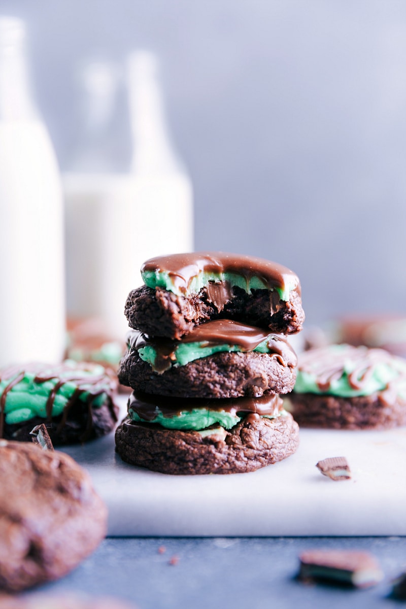 Delicious and festive Mint Brownie Cookies -- like a mint brownie but in cookie form! The chocolate cookie base is made with a brownie mix for an authentic brownie-tasting cookie. These fudgy and rich cookies are topped with peppermint frosting and a chocolate mint ganache, but they can also be simplified and made without those extra toppers. Recipe via ChelseasMessyApron.com #brownies #mintchocolate #mint #chocolate #brownie #brookie #ganache View of three Mint Brownie Cookies stacked, with a bite taken out of the top one.