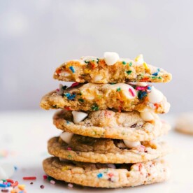 A stack of cake batter cookies, with one cookie broken open to reveal its delicious interior, and all of them topped with colorful sprinkles for a delightful treat.