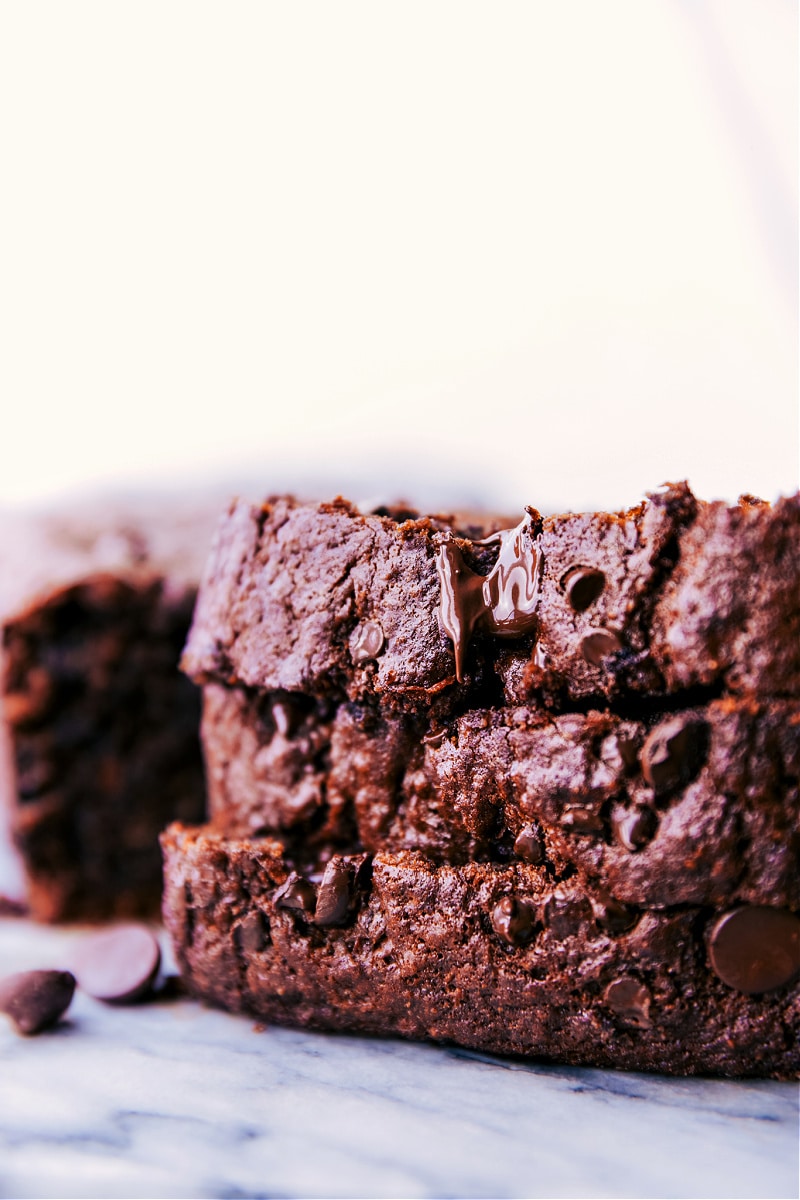 This tasty Chocolate Banana Bread is moist, deeply chocolate, and rich! We make this bread in two bowls -- no stand or hand mixers required. This banana bread also utilizes better-for-you ingredients -- less sugar than most banana breads, Greek yogurt, dark chocolate, coconut oil, blended up oats, and bananas of course! Recipe via chelseasmessyapron #breakfast #chocolatechips #dessert #best #quick #easy #simple #chocolate #banana #bread Image of the chocolate banana bread slices stacked on top of each other