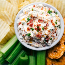 Baked potato dip creamy and topped with herbs and bacon surrounded by potato chips and celery sticks.
