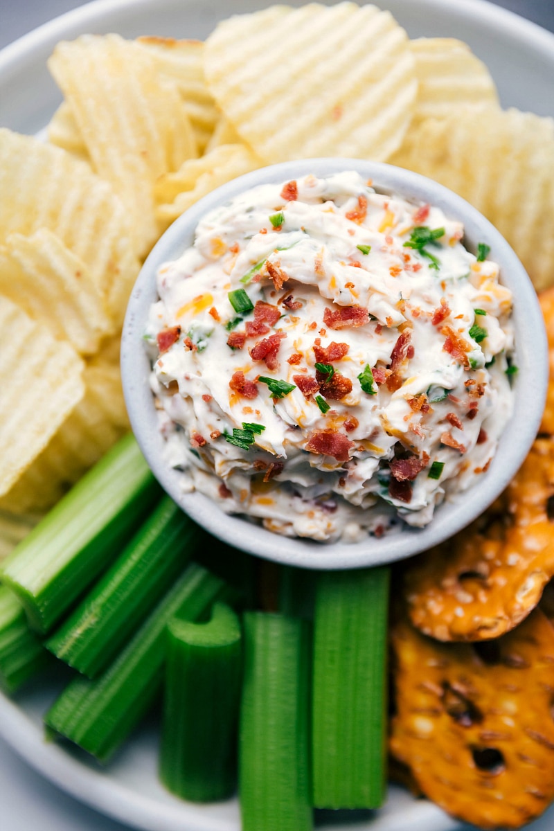 Baked potato dip creamy and topped with herbs and bacon surrounded by potato chips and celery sticks.