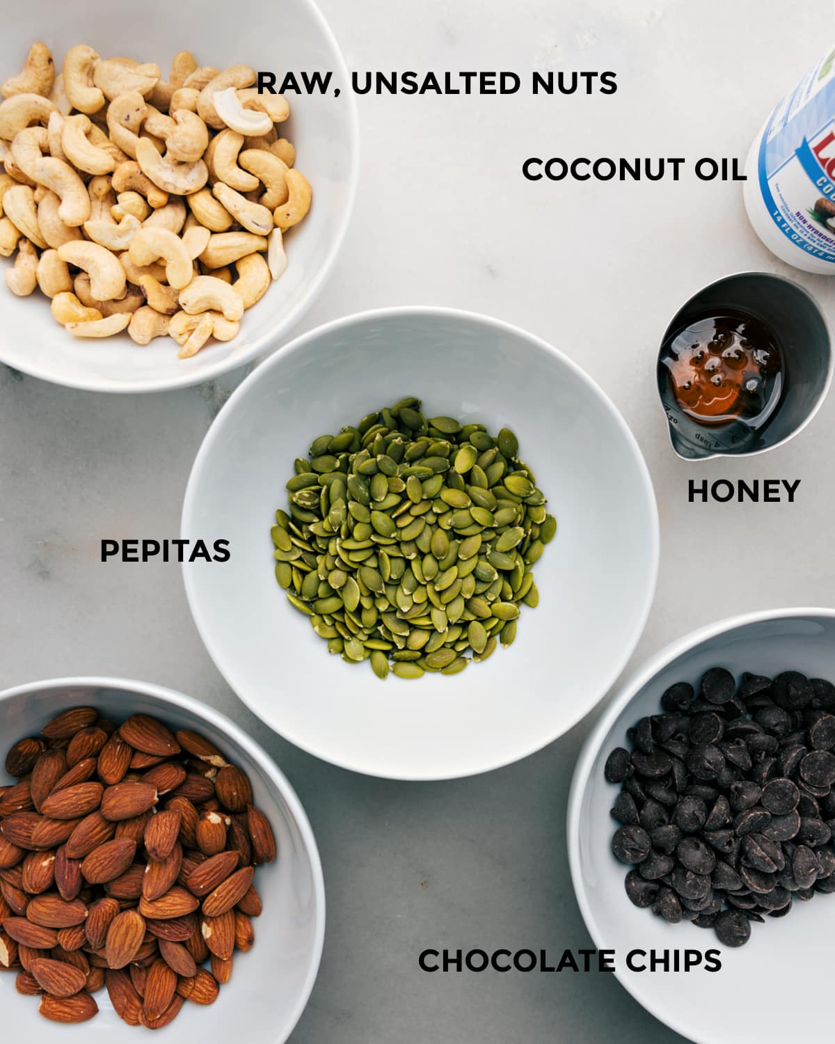 Visi šio recepto ingredientai, tokie kaip riešutai, kokosų aliejus, pepitai, medus ir šokolado drožlės, paruošti, kad būtų lengva surinkti.