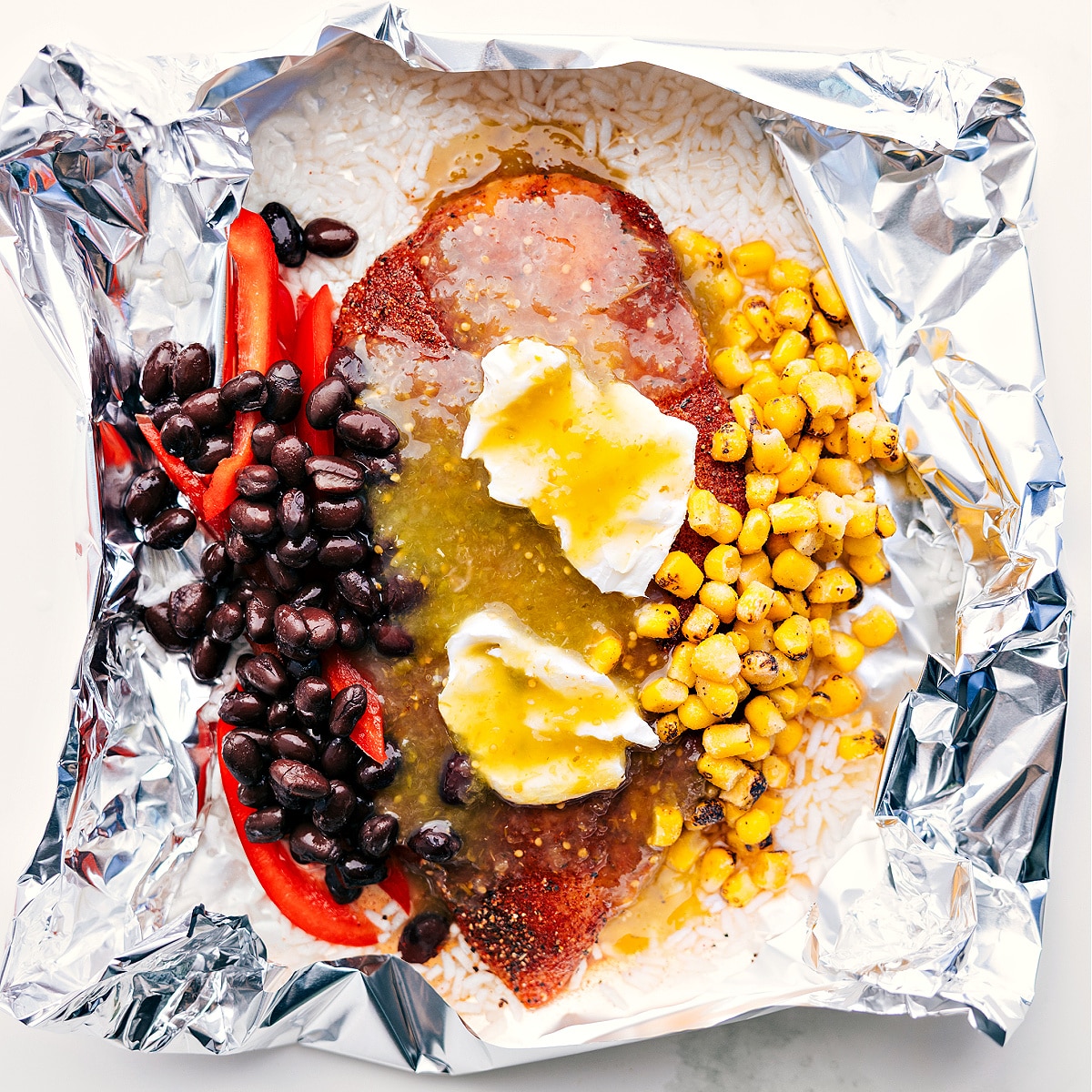 Foil packet filled with rice, corn, beans, cream cheese, salsa verde, and chicken, ready to be cooked over a fire, grill, or in the oven.