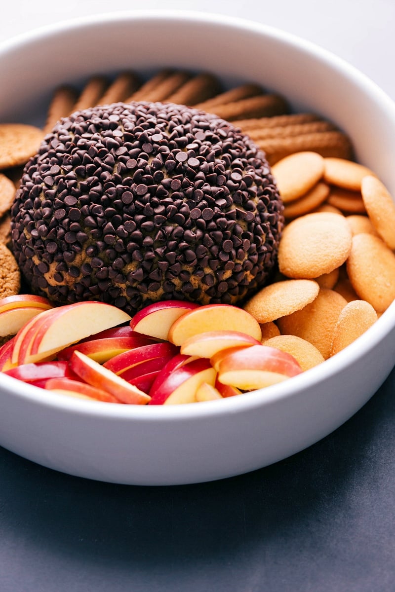 A delicious no bake pumpkin cheesecake ball presented in a bowl, surrounded by an assortment of dippers including vanilla wafers, apple slices, and more, offering a delightful and indulgent dessert spread.