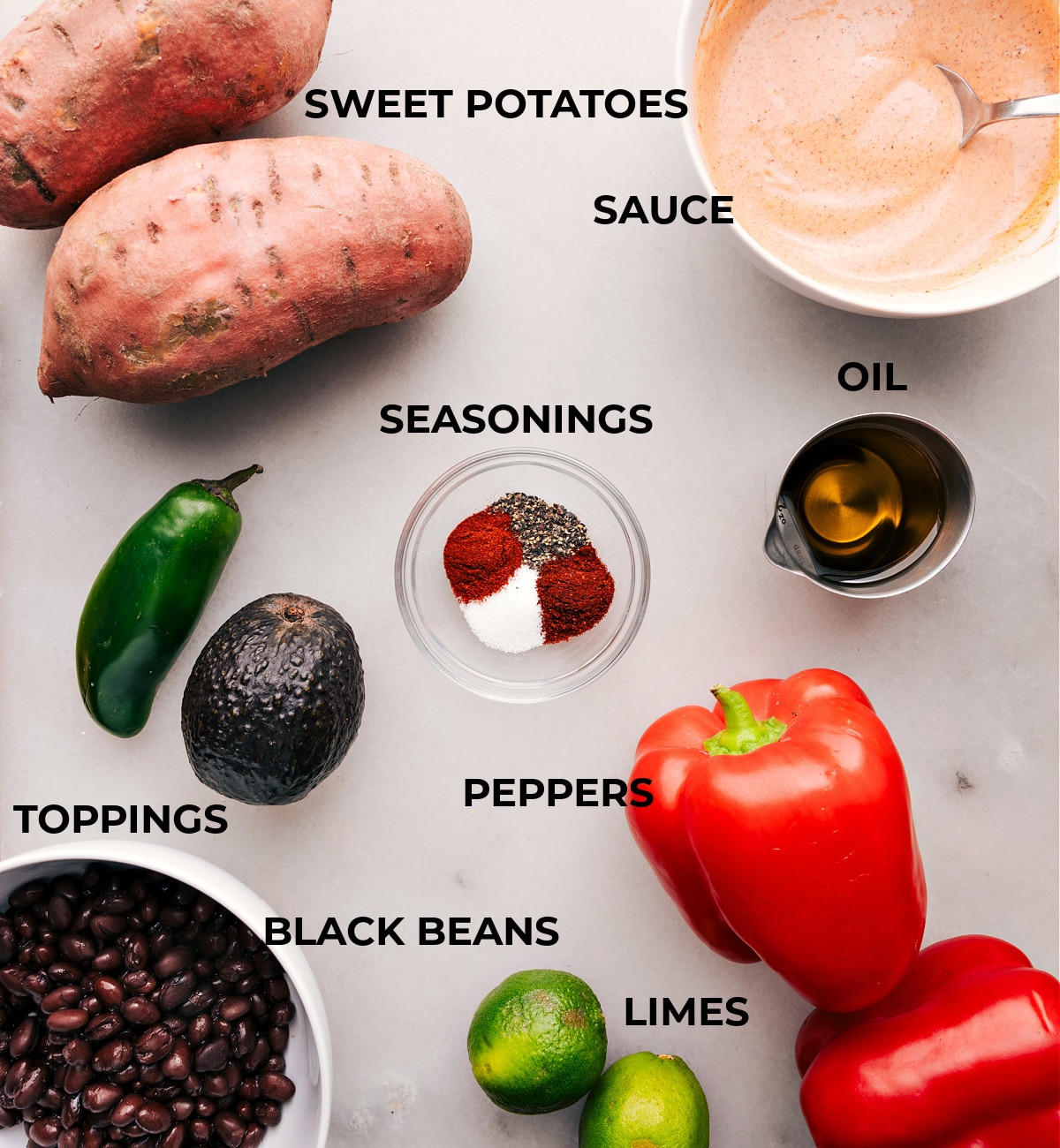 Ingredients for Sweet Potato and Black Bean Burrito Bowls including diced sweet potatoes, canned black beans, sliced peppers, halved avocado, and sliced jalapenos, halved limes, and a bowl of dressing.
