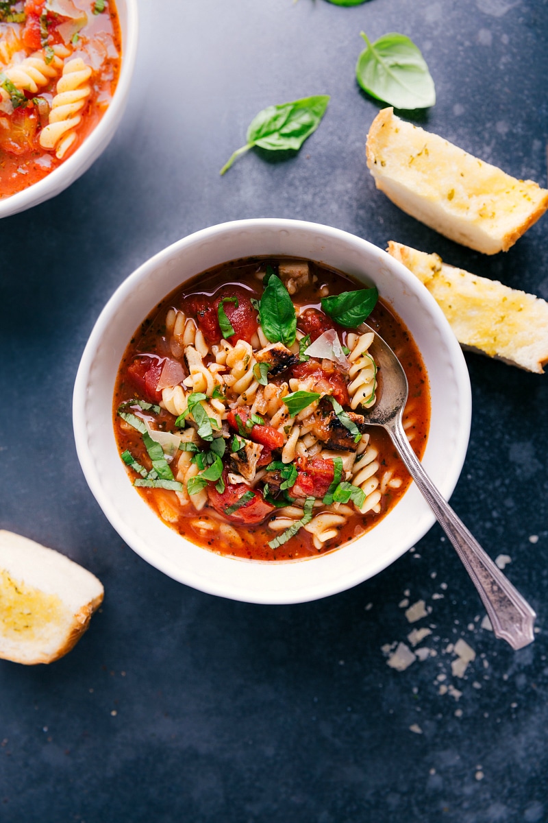 Overhead view of a bowl of Chicken Parmesan Soup, surrounded by buttered garlic bread, basil leaves and a few shreds of Parmesan cheese.