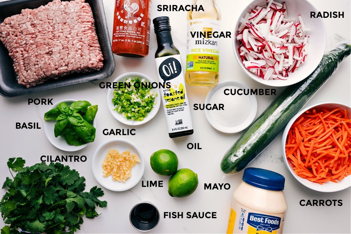 All the ingredients for this recipe prepped and ready for easy assembly, including pork, sriracha, vinegar, oil, green onions, basil, cilantro, garlic, lime, mayo, fish sauce, carrots, cucumber, sugar, radishes, and vinegar.