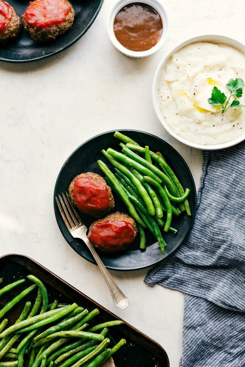 Overhead image of two mini meatloafs on a plate with green beans and a separate bowl of potatoes and gravy