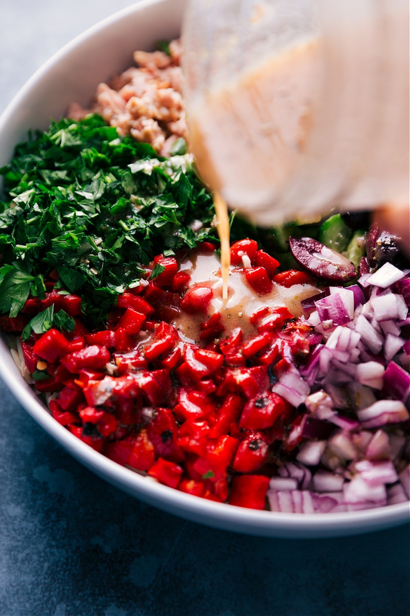 Our favorite Mediterranean Tuna Salad has tender orzo pasta, olive-oil-packed tuna, crisp cucumbers, smoky red peppers, tangy red onions, fresh herbs, and a lemon-oregano vinaigrette. Recipe via ChelseasMessyApron.com #tunasalad #tuna #vinaigrette #mediterranean #orzo #healthy #refreshing View of dressing being drizzled on the salad on this Mediterranean Tuna Salad