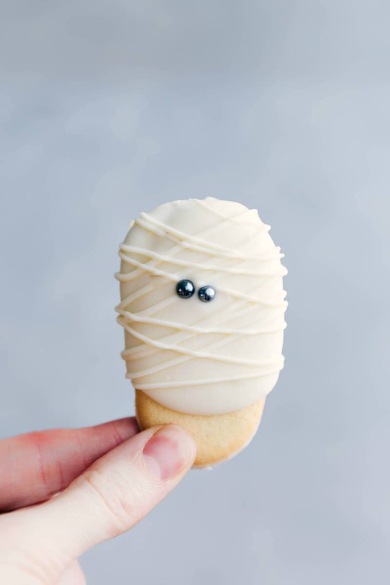 An adorable and festive mummy cookie being held, decorated with white icing and eyes to resemble a mummy, perfect for Halloween.