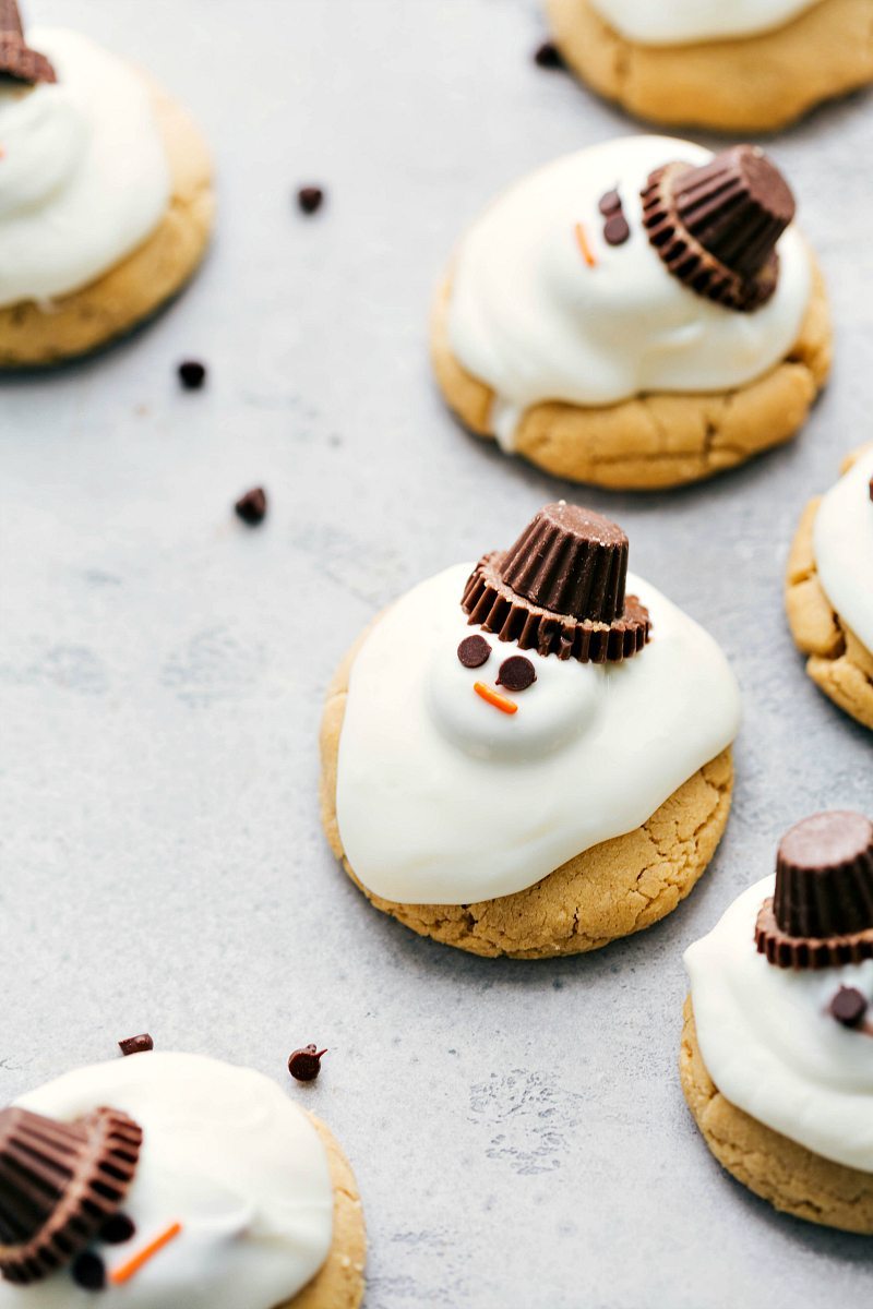 Adorable AND delicious easy-to-make melted snowman cookies. The cookie base is a peanut butter cookie and is topped with peanut butter cups and a white chocolate coating. Photo of several melted snowman cookies