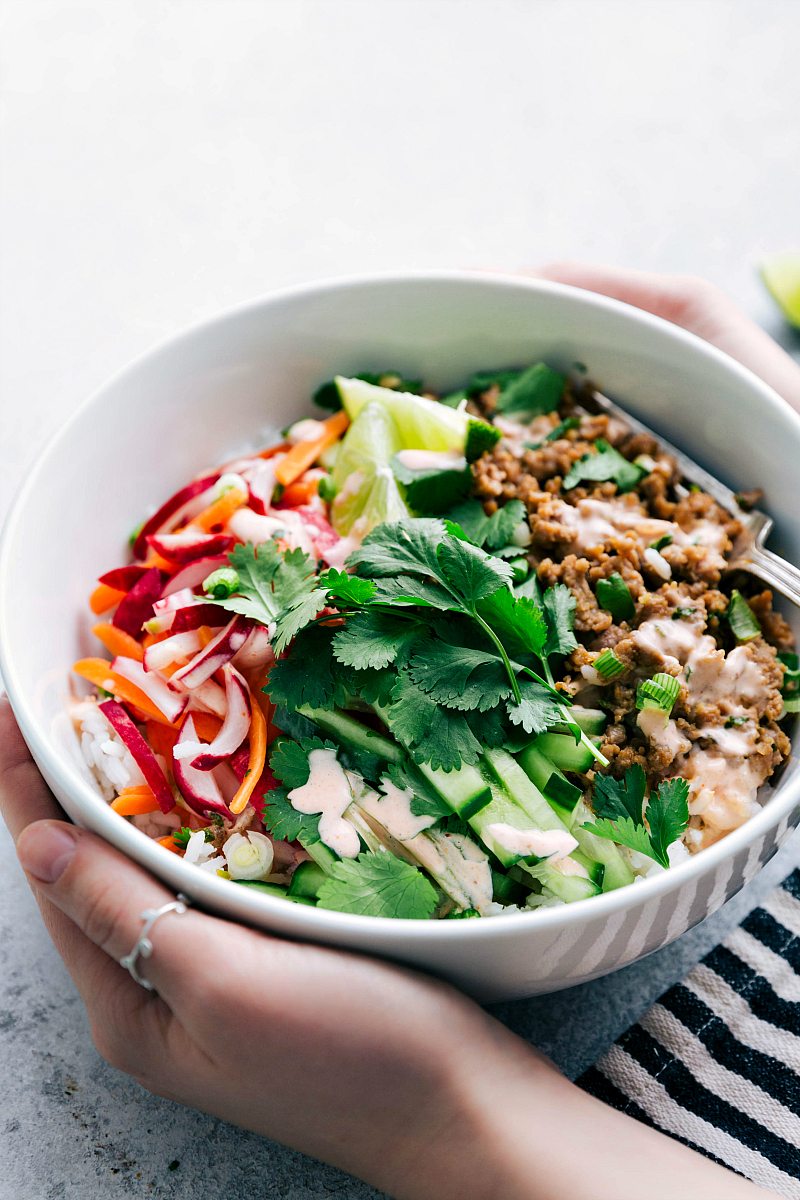 Side angle of white bowl holding banh mi ingredients: rice, ground pork mixture, cilantro, cucumber, lime, pickled radish and carrot, and Sriracha mayo