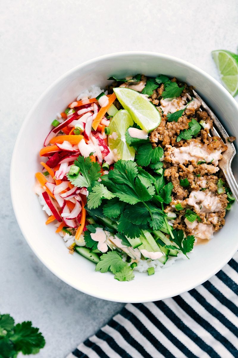 Picture of white bowl holding banh mi ingredients: rice, ground pork mixture, cilantro, cucumber, lime, pickled radish and carrot, and Sriracha mayo