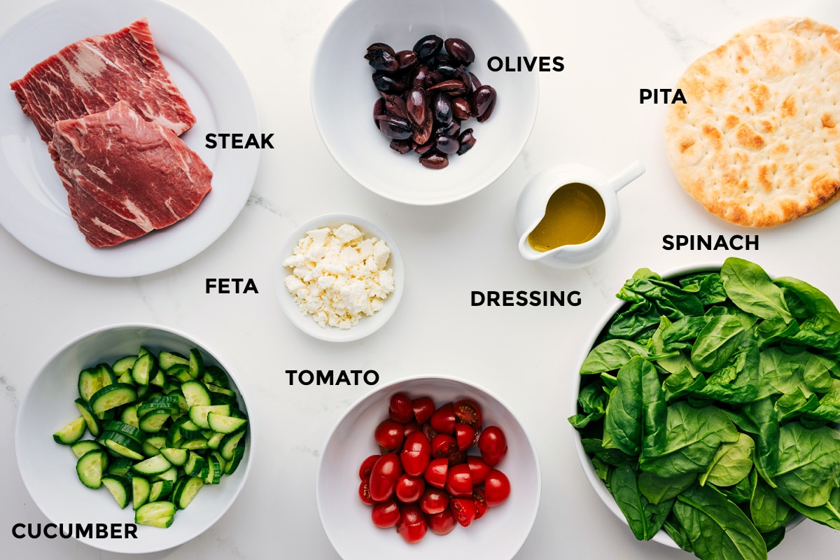 All the ingredients prepped for easy assembly, including sliced steak, olives, pita wedges, spinach, honey-lemon dressing, crumbled feta, cherry tomatoes, and cucumber.