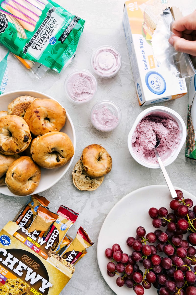 Overhead view of components of a healthy school lunch: bagels, string cheese, cream cheese, grapes and granola bars.