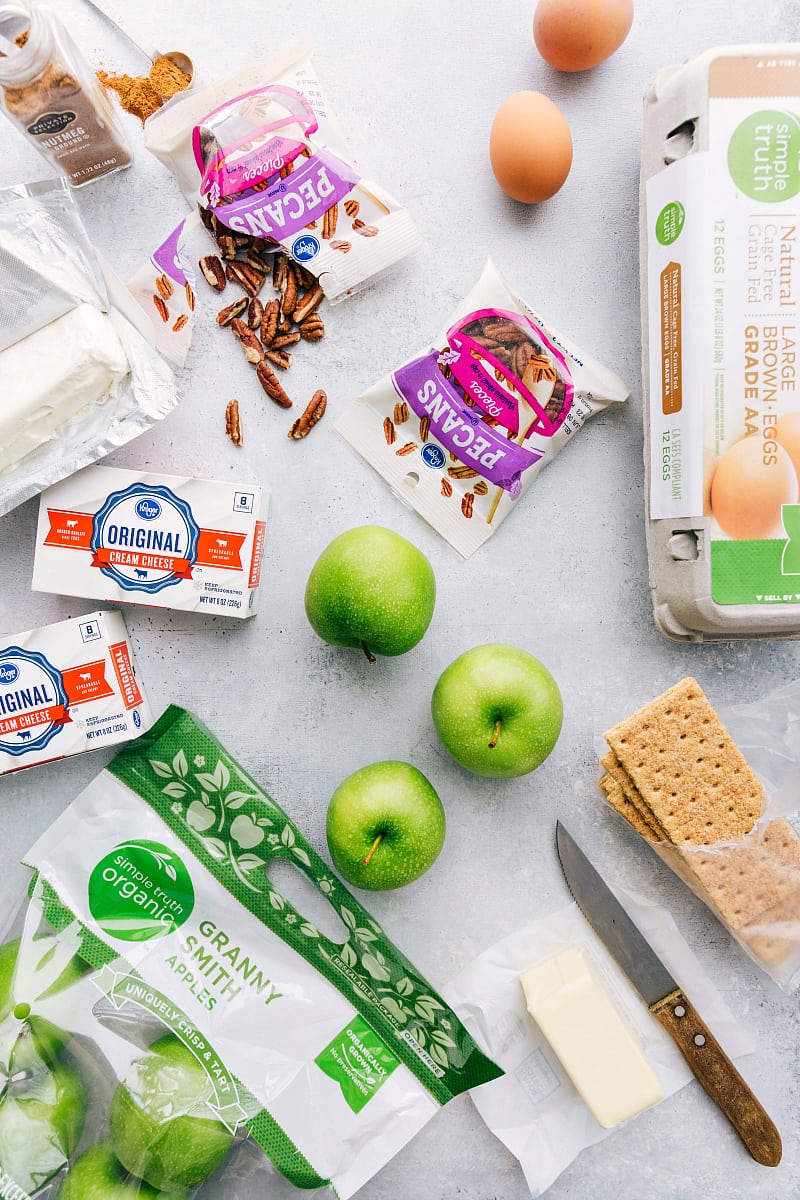 Various ingredients for apple caramel cheesecake bars, including apples, graham crackers, cream cheese, and more.