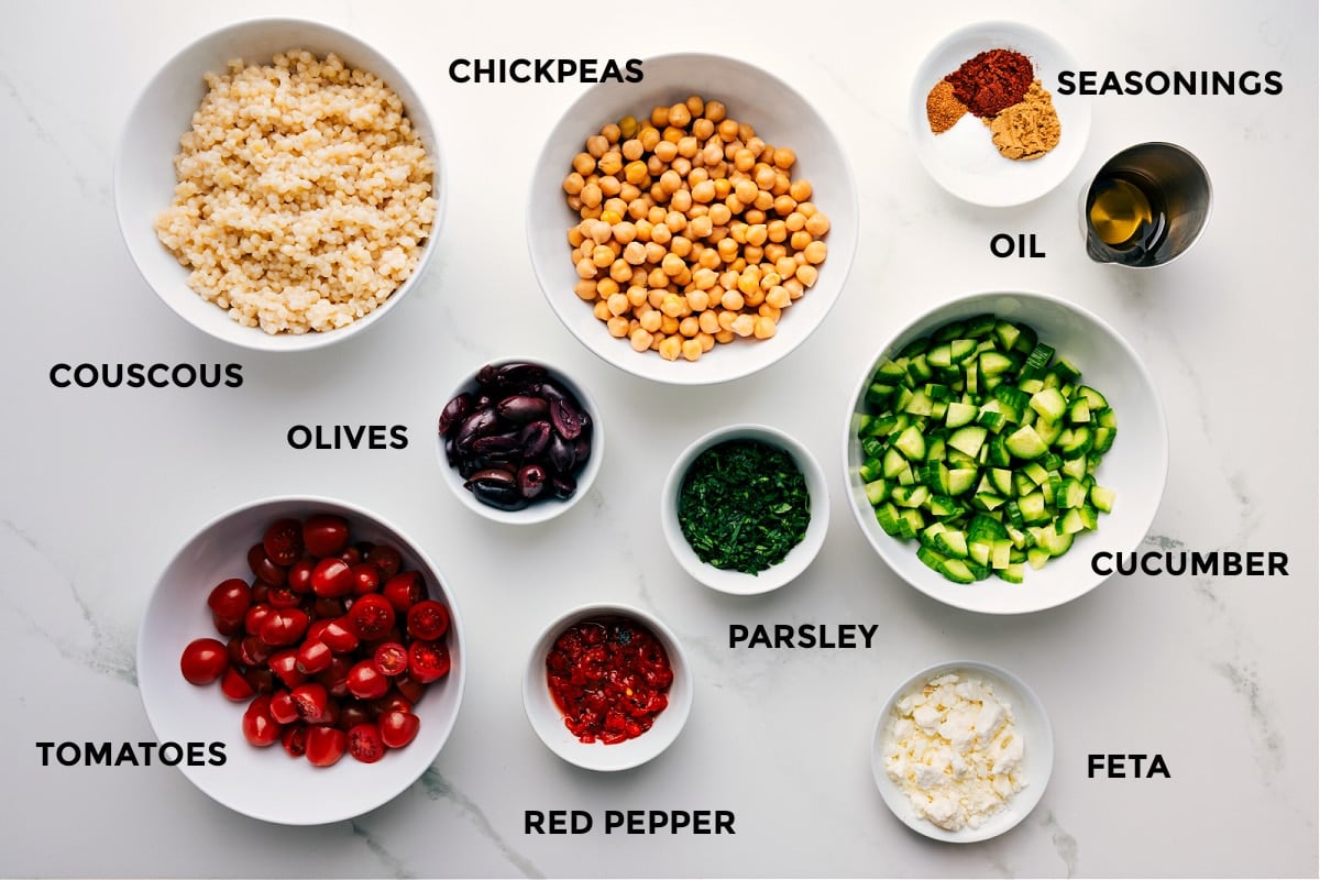 All the ingredients prepped for easy assembly, including chickpeas, couscous, seasonings, oil, cucumbers, parsley, feta, peppers, tomatoes, and olives.