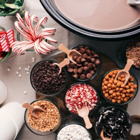 Assorted hot chocolate ingredients and toppings displayed on a table, including cups, chocolate shavings, whipped cream, and marshmallows.