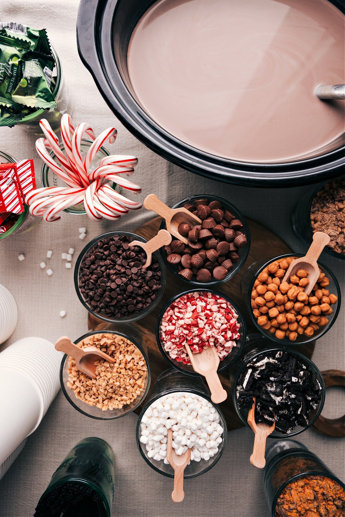 Assorted hot chocolate ingredients and toppings displayed on a table, including cups, chocolate shavings, whipped cream, and marshmallows.