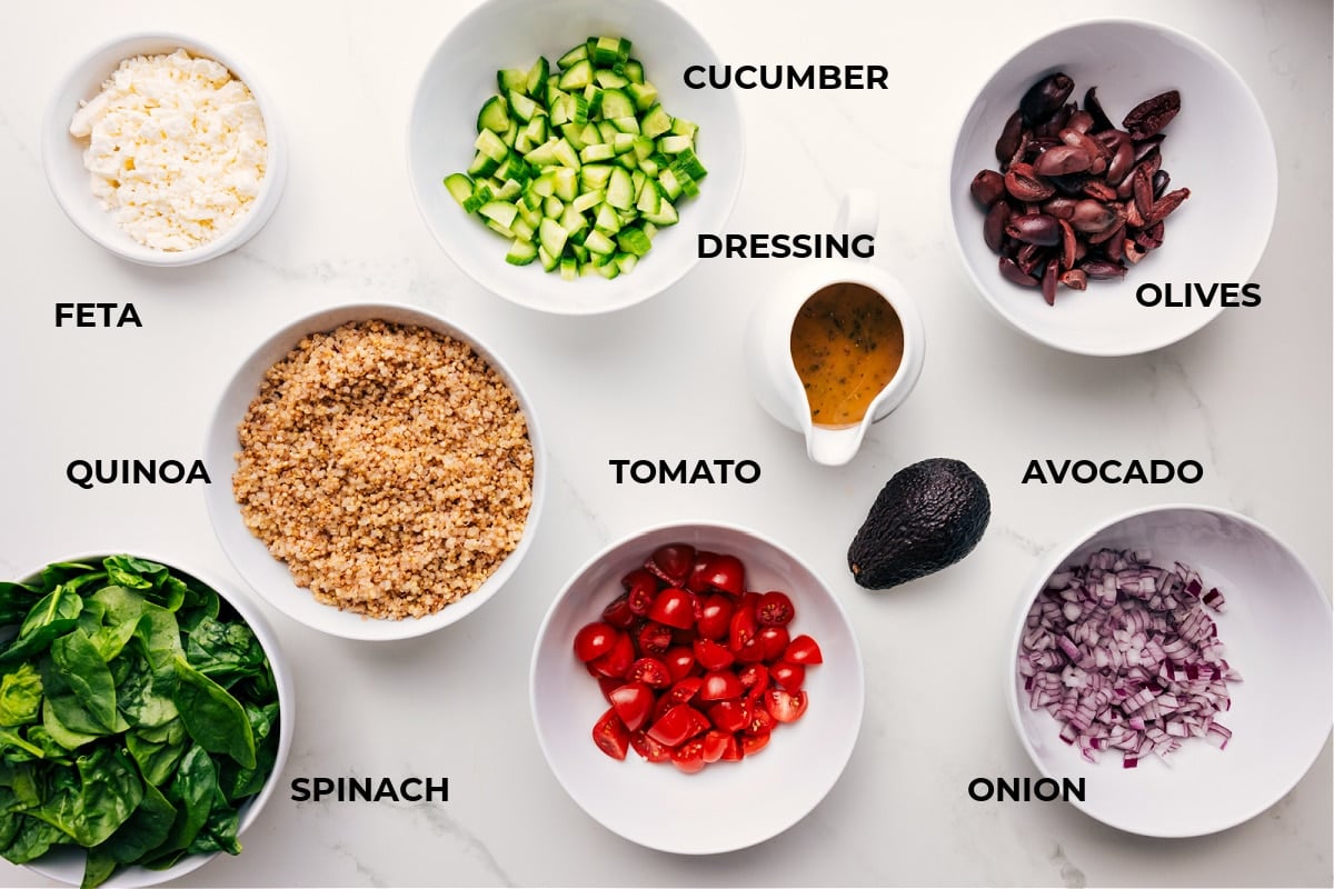 Ingredients in this recipe prepped out for easy assembly including the feta, quinoa, spinach, cucumber, dressing, olive, avocado, onion, and tomato.