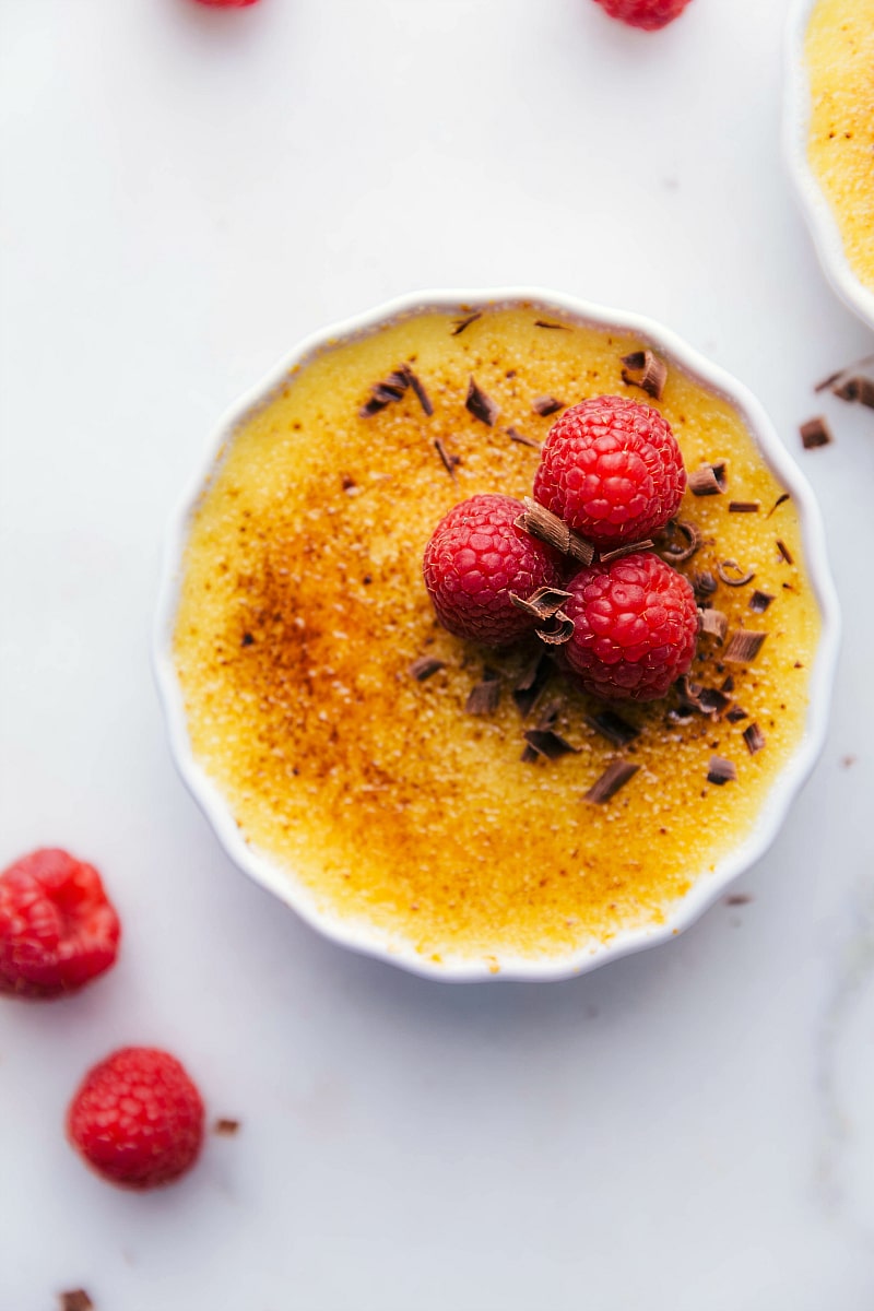 Tuxedo Crème Brûlée is a chocolatey twist on the traditional crème brûlée recipe. This dessert has a chocolate ganache bottom, creamy custard filling, and a caramelized sugar topping. Top these brulée with fresh berries and chocolate curls for a delicious show-stopping dessert. Recipe via ChelseasMessyApron.com #cremebrulee #tuxedo #ganache #French #dessert #elegant Overhead image of the Tuxedo Crème Brûlée garnished with chocolate curls and raspberries.