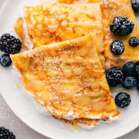 Plate of three crepes with berries and syrup drizzled on top.