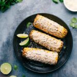 Ready-to-eat Elote on a plate with limes and cilantro around them.