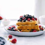 Stack of waffles with berries and whipped cream on top with syrup drizzling down.