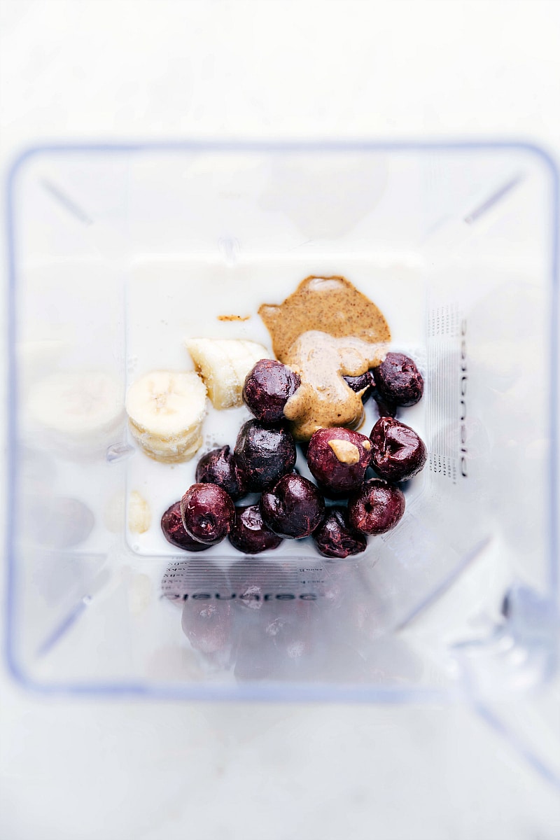 All the ingredients for a healthy cherry smoothie in the blender, including milk, cherries, almond butter, and frozen bananas, ready to be blended together for the healthy cherry smoothie recipe.