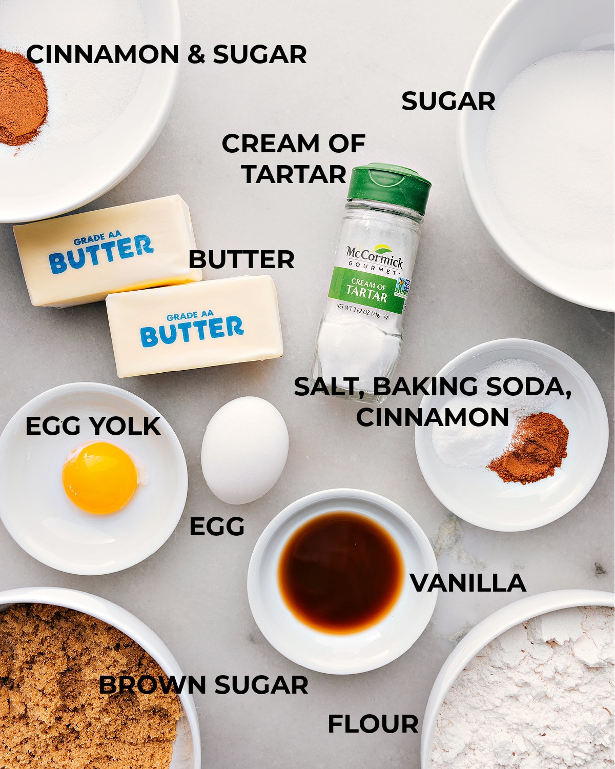 Various ingredients for making the snickerdoodle recipe arranged on a kitchen counter, including flour, sugar, eggs, and cinnamon.
