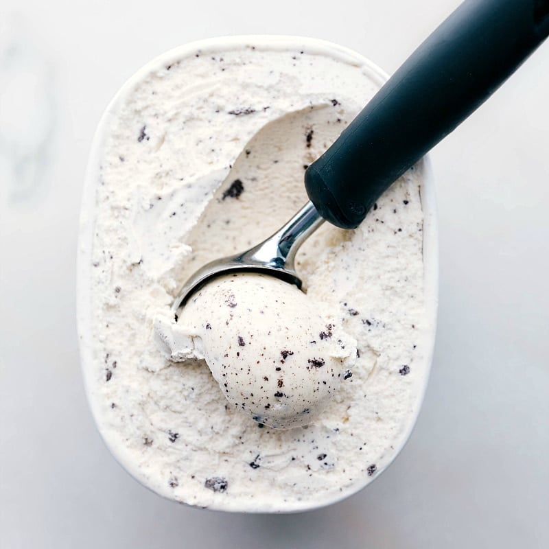 A container of cookies and cream ice cream with a scoop in it for this cake made with ice cream sandwiches.