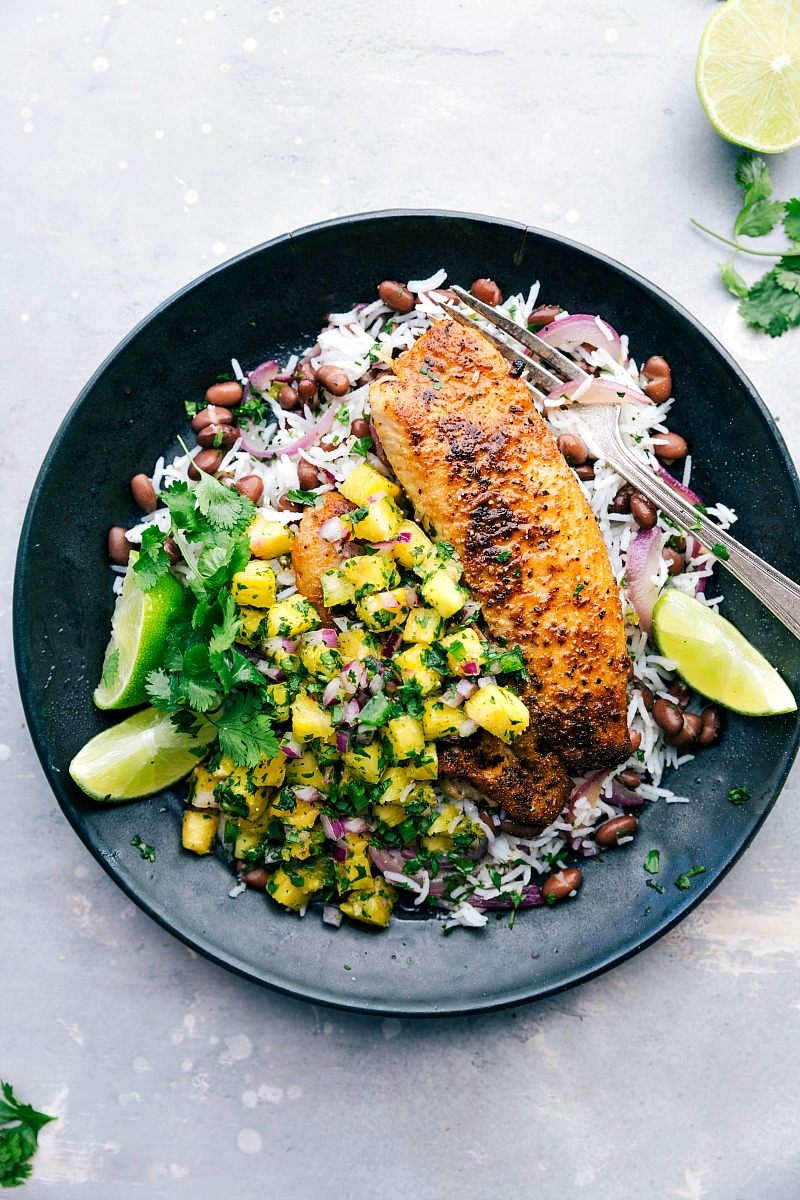 A plate featuring blackened tilapia served with pineapple salsa, rice, and black beans, presenting a flavor-packed and delicious meal.