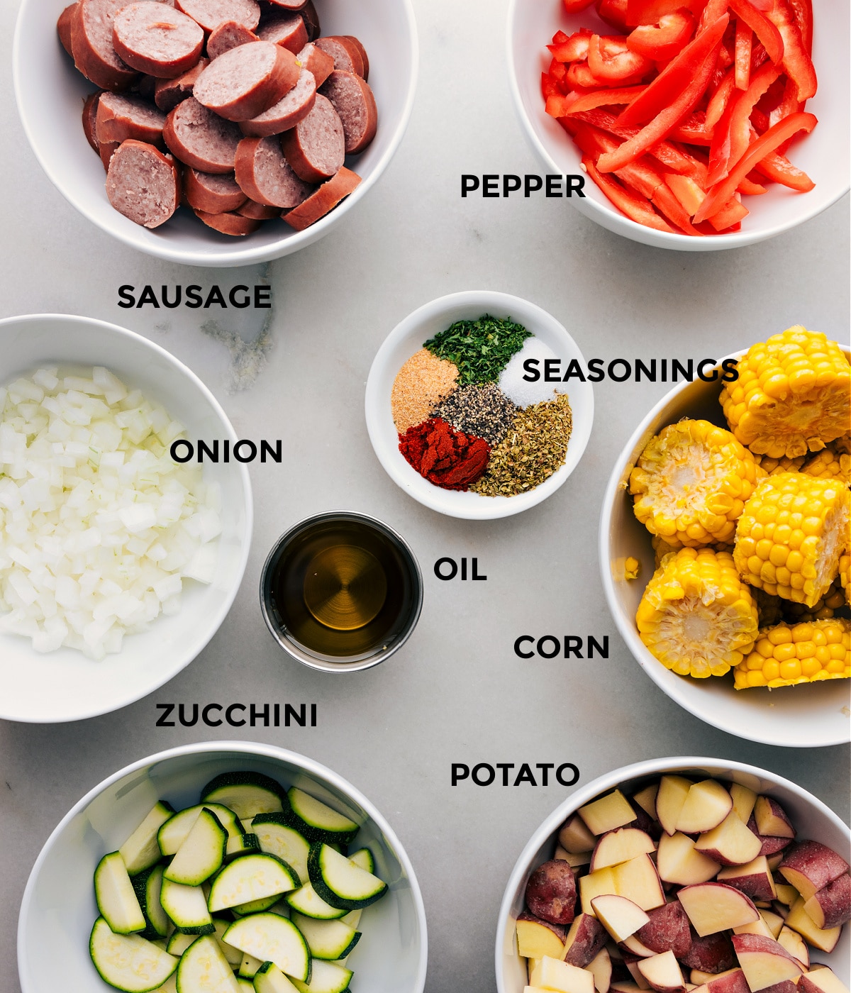 Visi šio recepto ingredientai, įskaitant dešrą, pipirus, svogūną, prieskonius, kukurūzus, cukinijas, bulves ir aliejų, paruošti, kad būtų lengva surinkti.