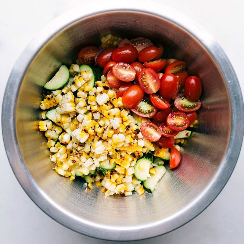 A bowl filled with the corn and zucchini salad, the addition of fresh tomatoes and more ingredients, capturing the moment before mixing to create a flavorful salad.