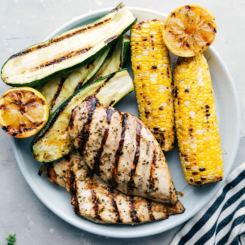 Overhead image of Greek Chicken with grilled zucchini, corn, and lemons on the side.