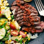 Sliced grilled flat iron steaks, generously topped with sun-dried tomato sauce, served with a side of grilled corn and zucchini salad, creating a hearty and delicious meal.