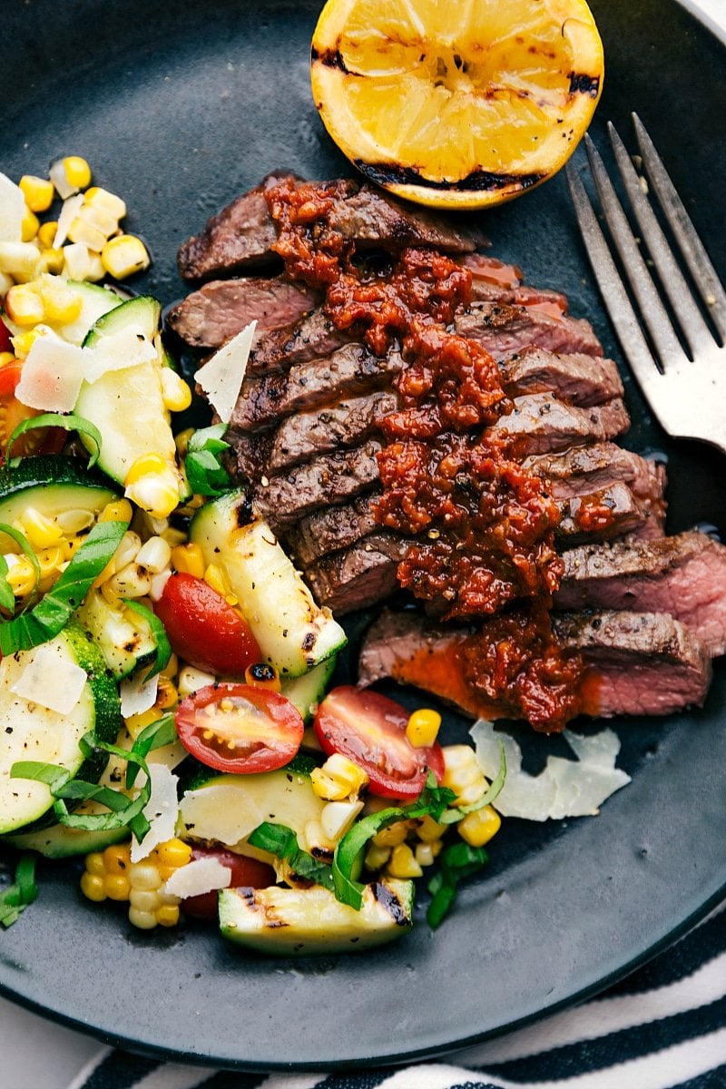 Sliced grilled flat iron steaks, generously topped with sun-dried tomato sauce, served with a side of grilled corn and zucchini salad, creating a hearty and delicious meal.