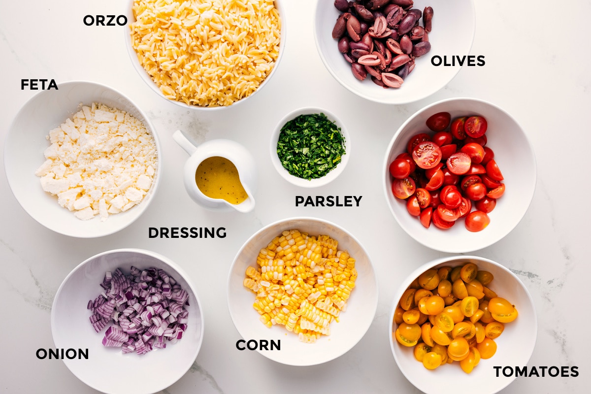 All the ingredients in this recipe prepped out for easy assembly including the feta, onion, dressing, corn, tomatoes, olives, parsley, and orzo.
