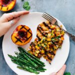 Peach salsa chicken served on a plate, accompanied by green beans and a freshly grilled peach on the side.