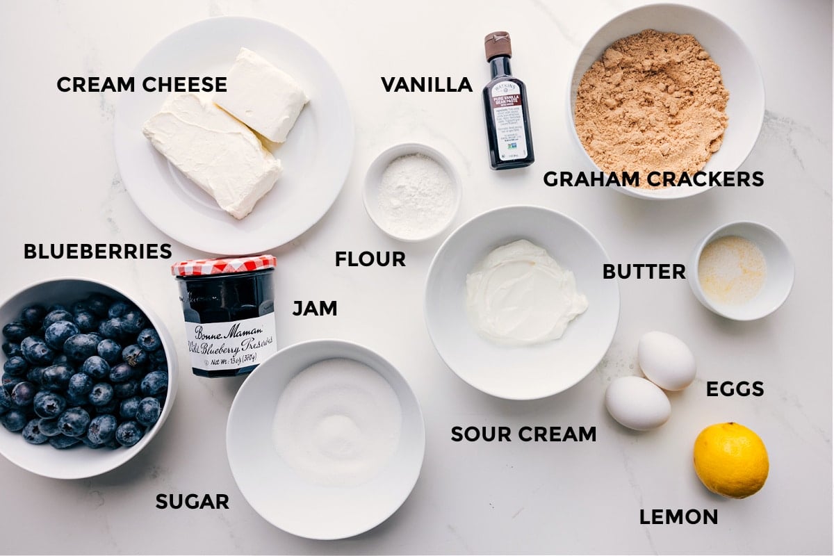 Ingredients In Mini Blueberry Cheesecakes All the ingredients in this recipe prepped out for easy assembly including the cream cheese, blueberries, jam, sugar, flour, sour cream, eggs, lemon, butter, graham crackers, and vanilla.