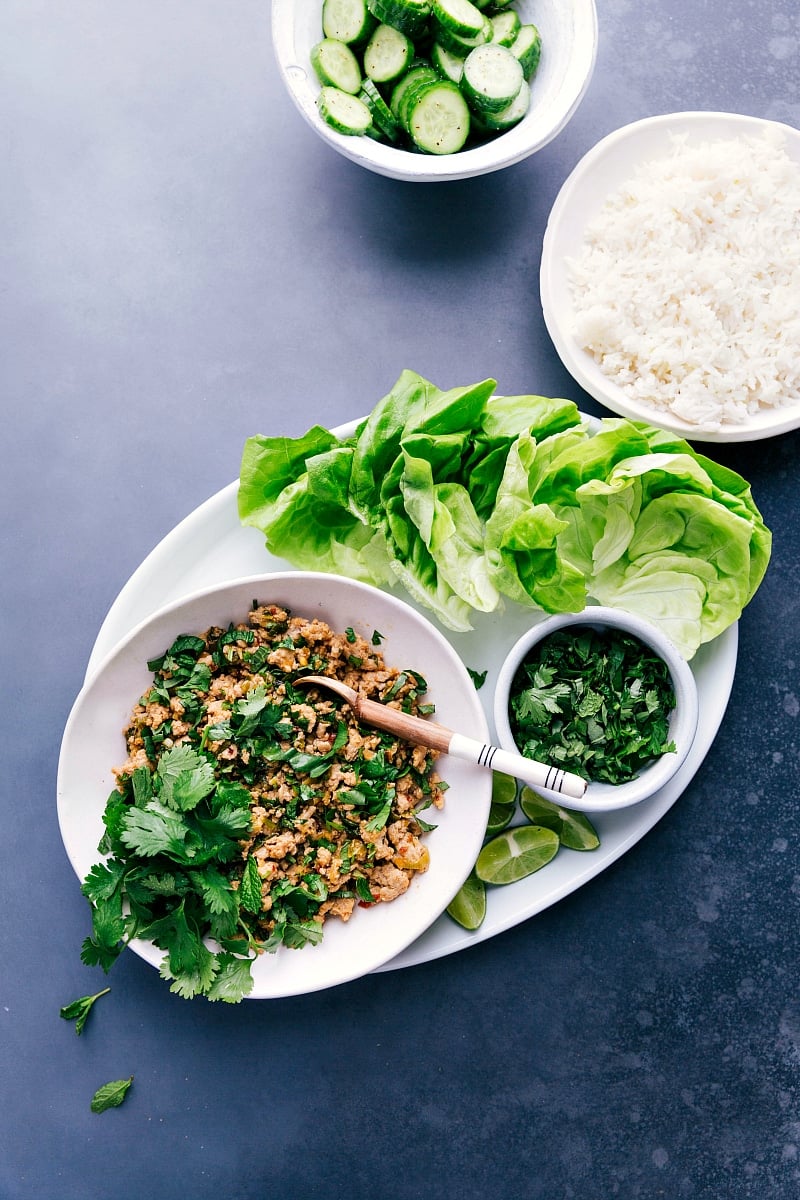 Overhead view of Chicken Larb on a platter, with a plate of rice and a bowl of sliced cucumbers to accompany.